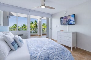 A bedroom with a bed, a dresser, a television, and a balcony with a view of palm trees. - Beachfront Penthouse with Private Terrace, Ocean & El Yunque Views – Casa Bacci (Río Grande)