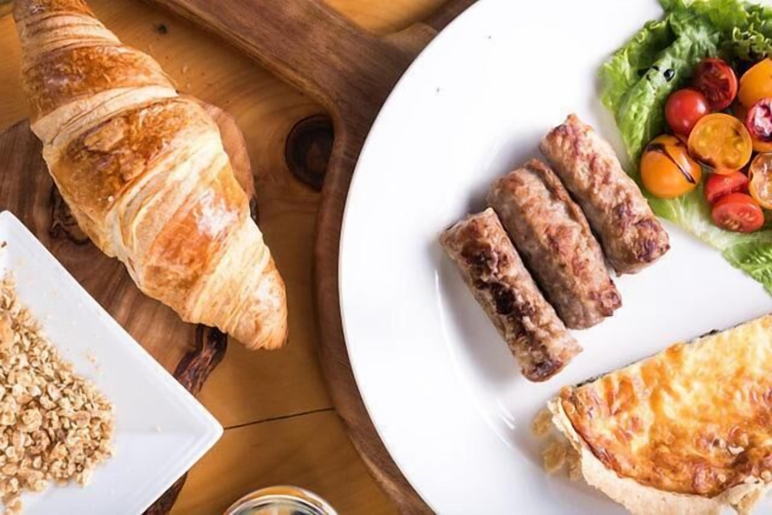 Una mesa de madera con un croissant, un plato con salchichas y una quiche, y un plato con ensalada.
