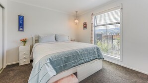Quarto com cama, mesa de cabeceira, vaso com flores, janela com persianas e luminária fixada na parede.