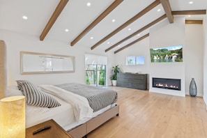 Un dormitorio moderno con cama, chimenea, televisor y un ventanal grande.