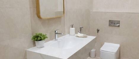 Un baño moderno con un espejo grande, un lavamanos blanco, una planta pequeña y un inodoro.
