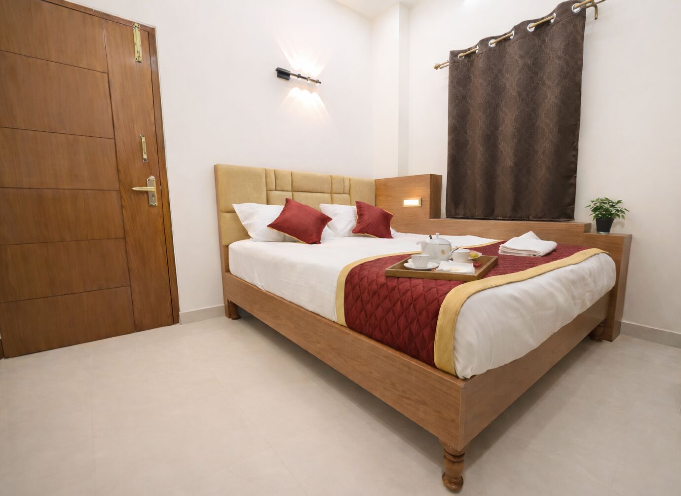 A hotel room with a wooden bed, a bedside table with a lamp, a window with curtains, and a door.