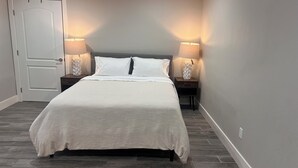 Una cama bien hecha con ropa de cama blanca en una habitación con paredes grises y suelo de madera oscura. Hay dos mesitas de noche con lámparas a cada lado de la cama.