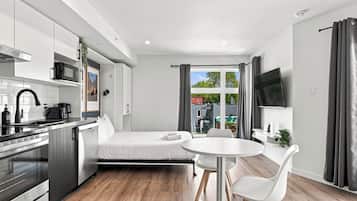 A compact hotel room with a kitchenette, a bed, a dining area with two chairs, a TV mounted on the wall, and a view of the outside through a large window.