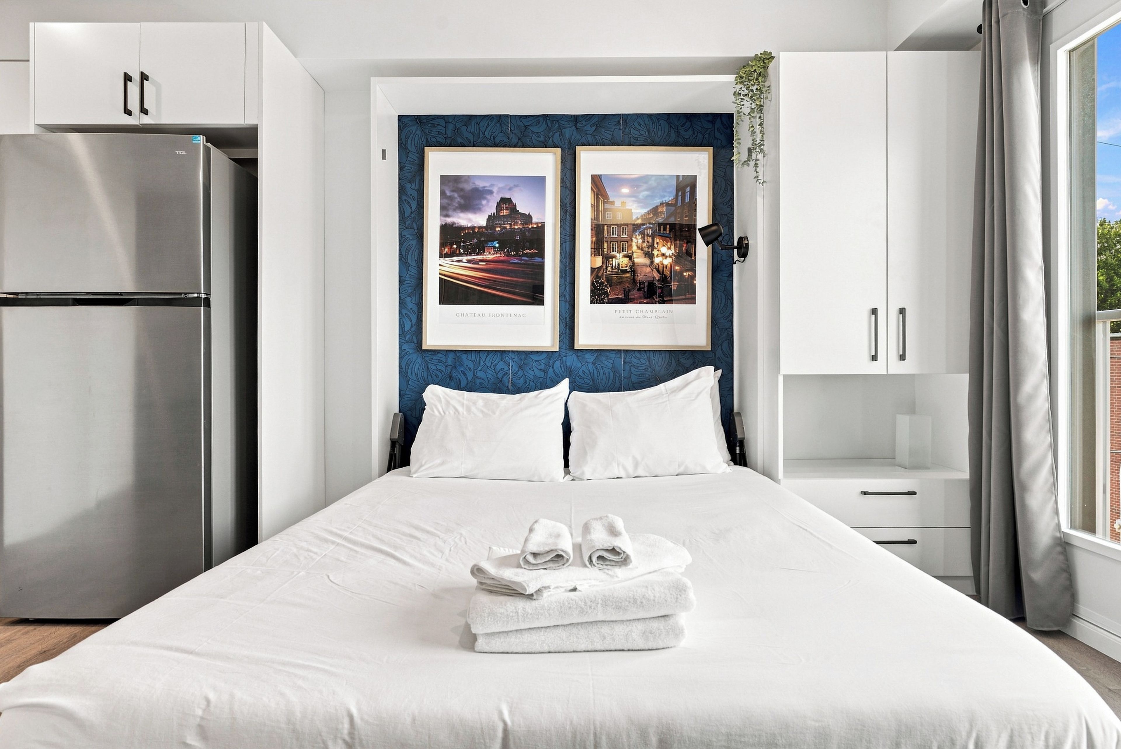 A hotel room with a bed, a minibar, a window with curtains, and two framed pictures on the wall.