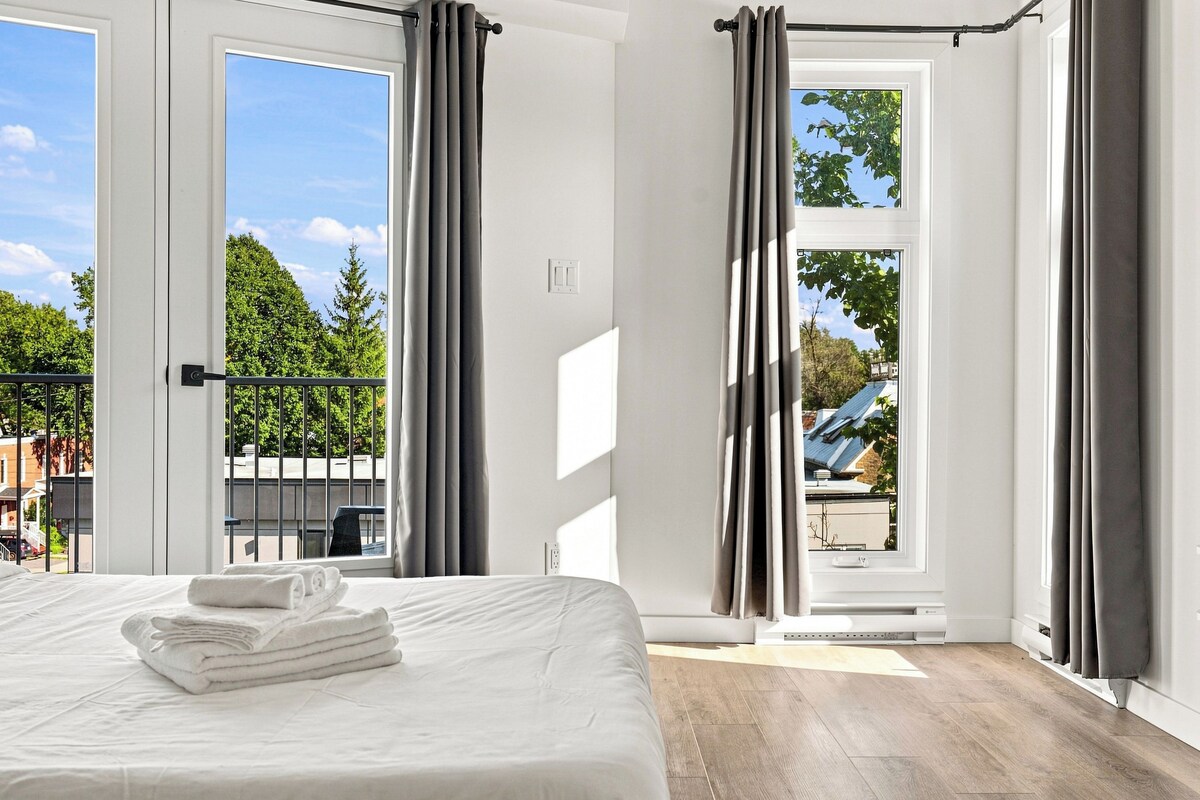 A hotel room with a bed, a window with curtains, a view of trees, and a balcony.