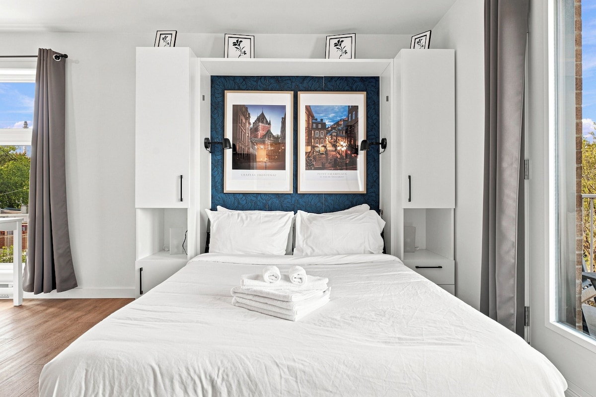 A hotel room with a large bed, white bedding, a view of the city, and two framed pictures on the wall.