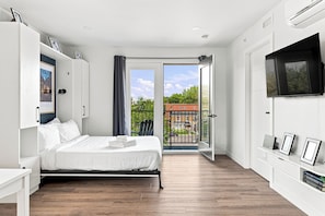 A modern bedroom with a bed, a TV, and a balcony with a view of buildings.