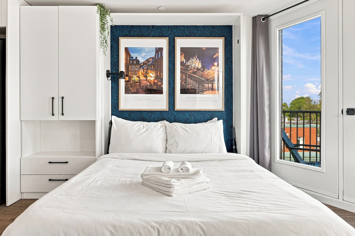 A bedroom with a bed, white bedding, two framed pictures of Parisian landmarks, a window with a view, and a plant on the wall.