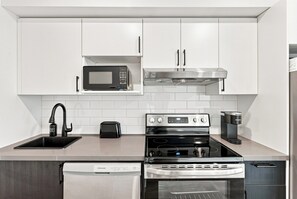 A modern kitchen with white cabinetry, a built-in microwave, a stainless steel range hood, a toaster, an oven, and a dishwasher. - Appart'hôtel Viveo - Par Aneyro (Québec)
