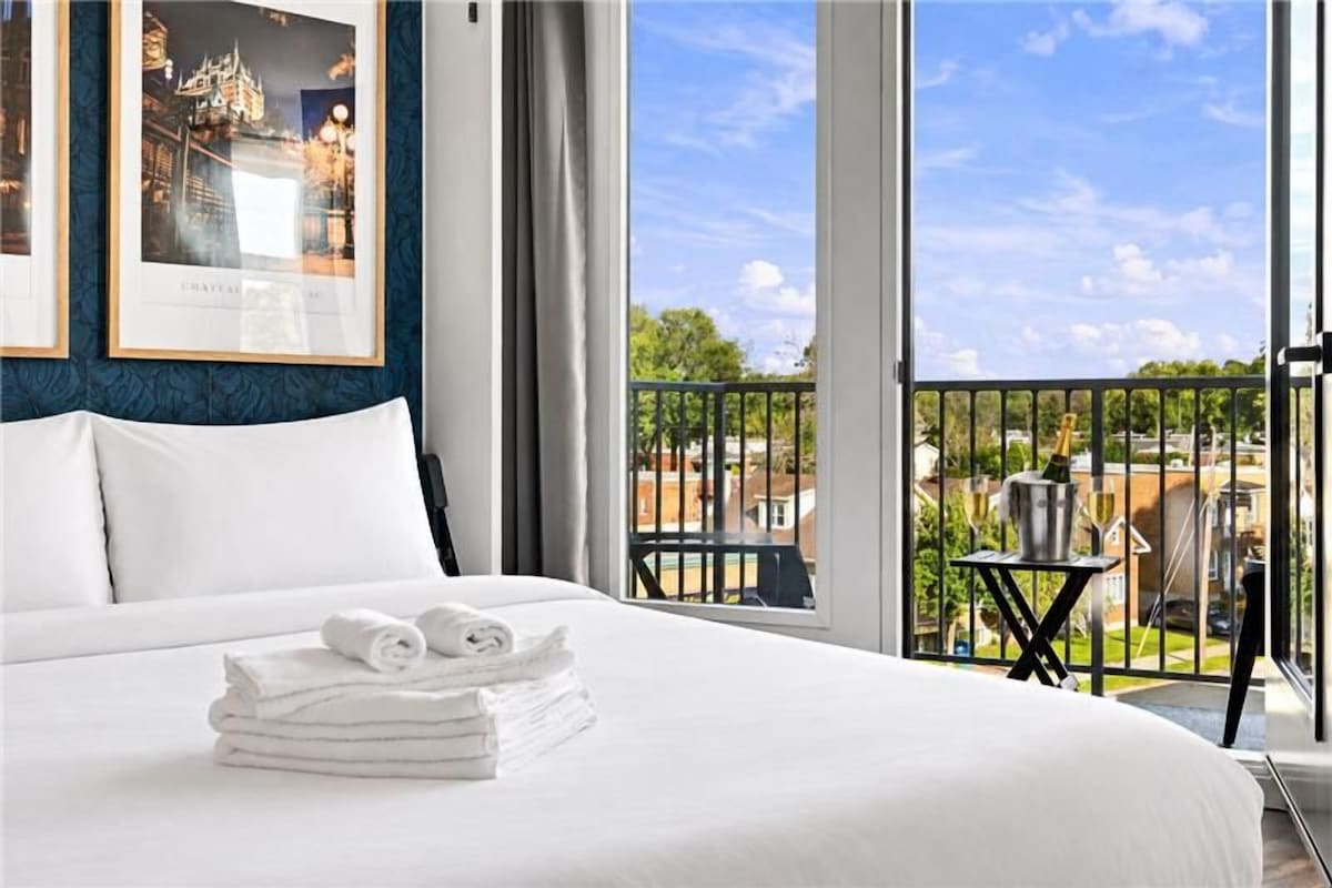A hotel room with a large bed, a view of the city, and a balcony with a table and chair.