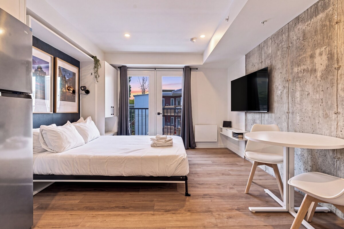 A modern hotel room with a bed, a desk, a chair, a flat-screen TV, and a balcony with a view.