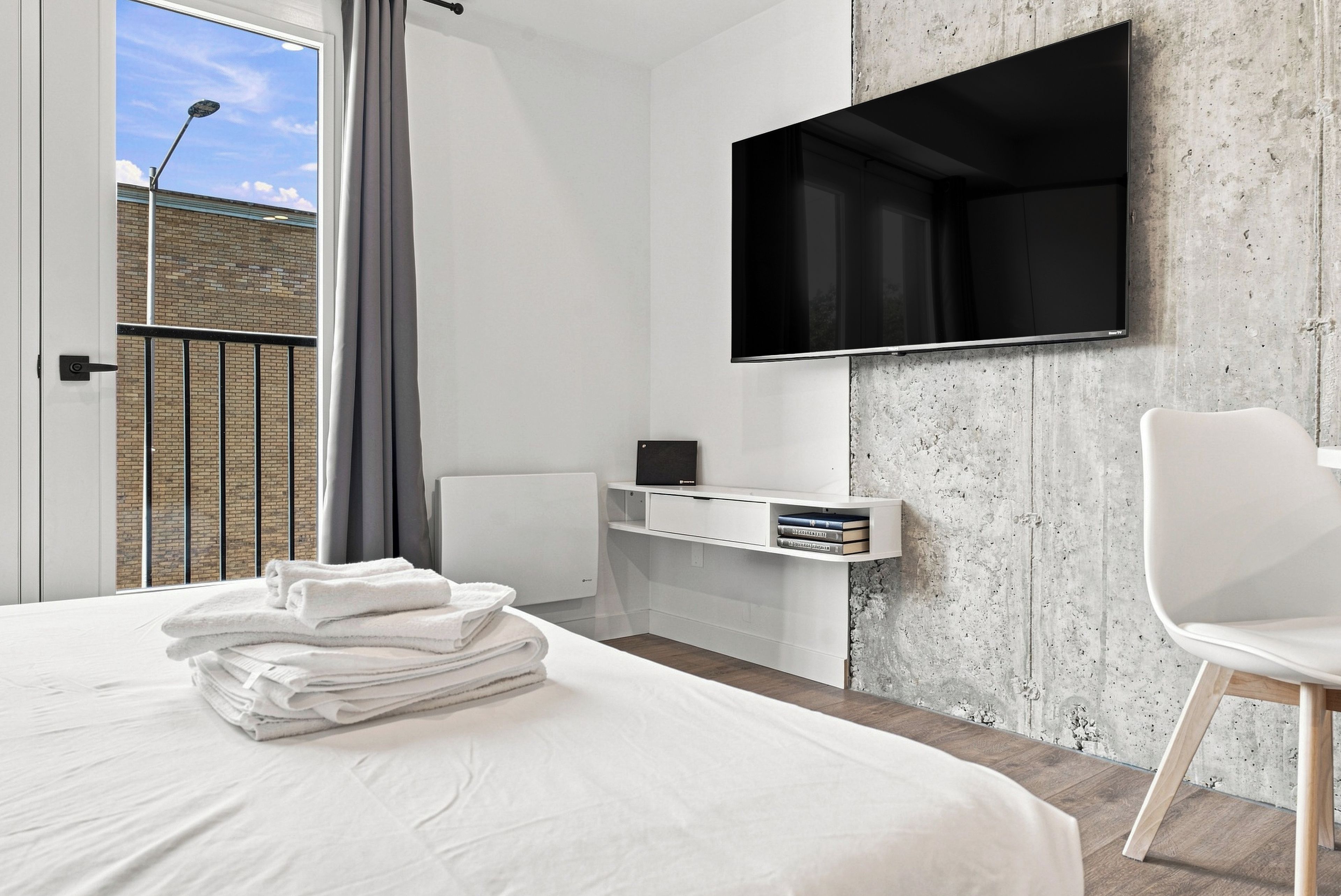 A modern hotel room with a large bed, a flat-screen TV mounted on a textured wall, a small desk with a chair, and a view of a building through a window with a curtain.