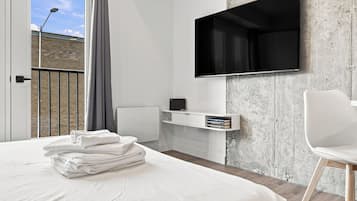 A modern hotel room with a large bed, a flat-screen TV mounted on a textured wall, a small desk with a chair, and a view of a building through a window with a curtain.