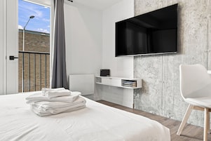 Une chambre d’hôtel moderne avec un grand lit, un téléviseur à écran plat fixé à un mur texturé, un petit bureau avec une chaise et une vue sur un immeuble à travers une fenêtre avec un rideau.