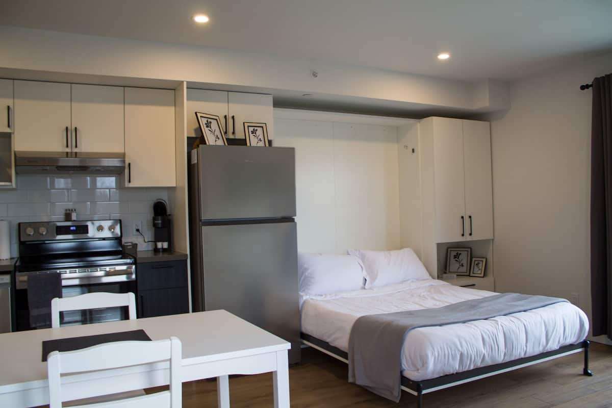 A compact hotel room with a kitchenette, a bed, a small table, and a chair.
