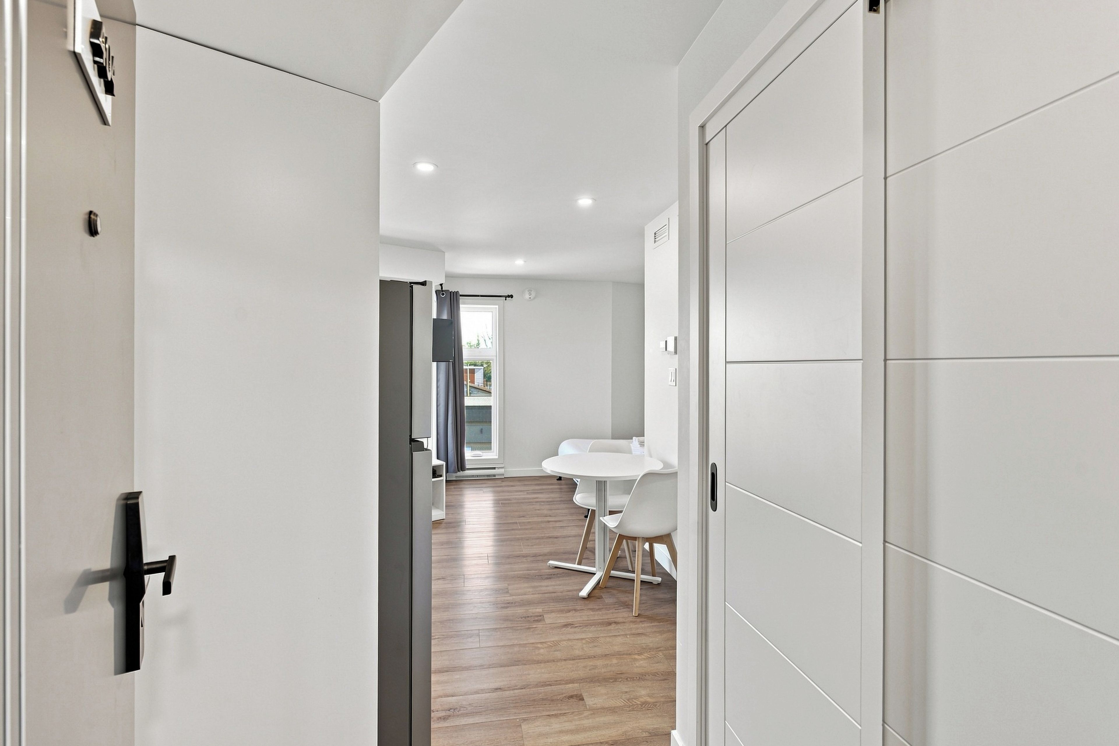 Un pasillo que conduce a un comedor con una mesa y sillas, una puerta a la izquierda y una pared blanca a la derecha.
