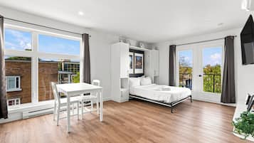Un dormitorio moderno con una cama, un escritorio y una silla. Hay ventanas amplias y un balcón.