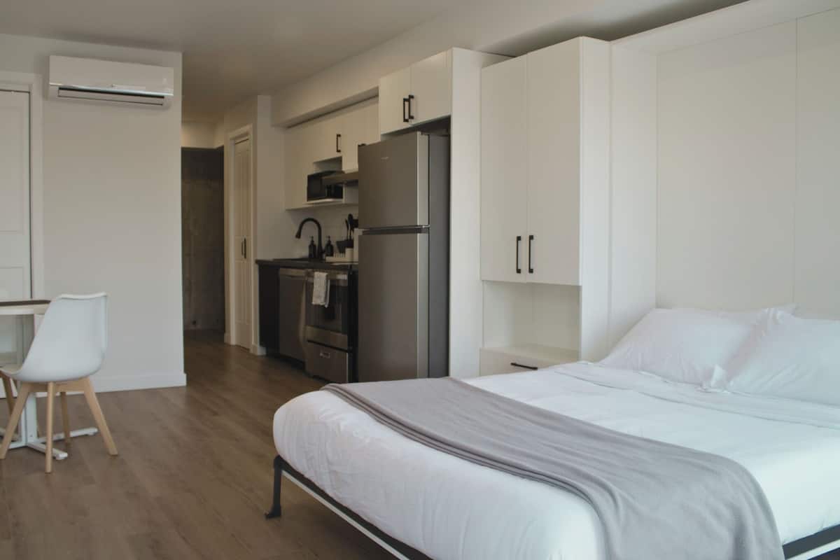 A modern bedroom with a bed, a desk with a chair, a kitchenette with a refrigerator, and an air conditioning unit.