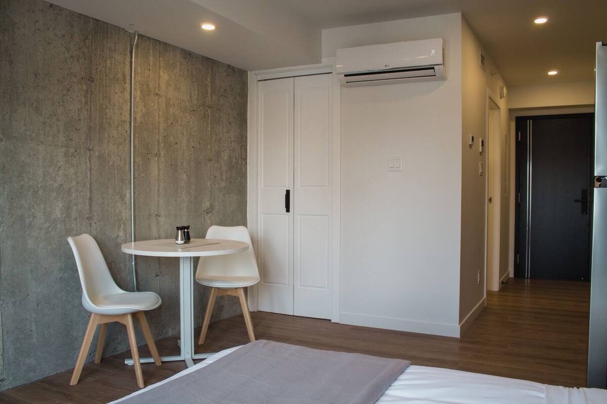 A room with a bed, two chairs, a small round table, a wardrobe, and an air conditioning unit.