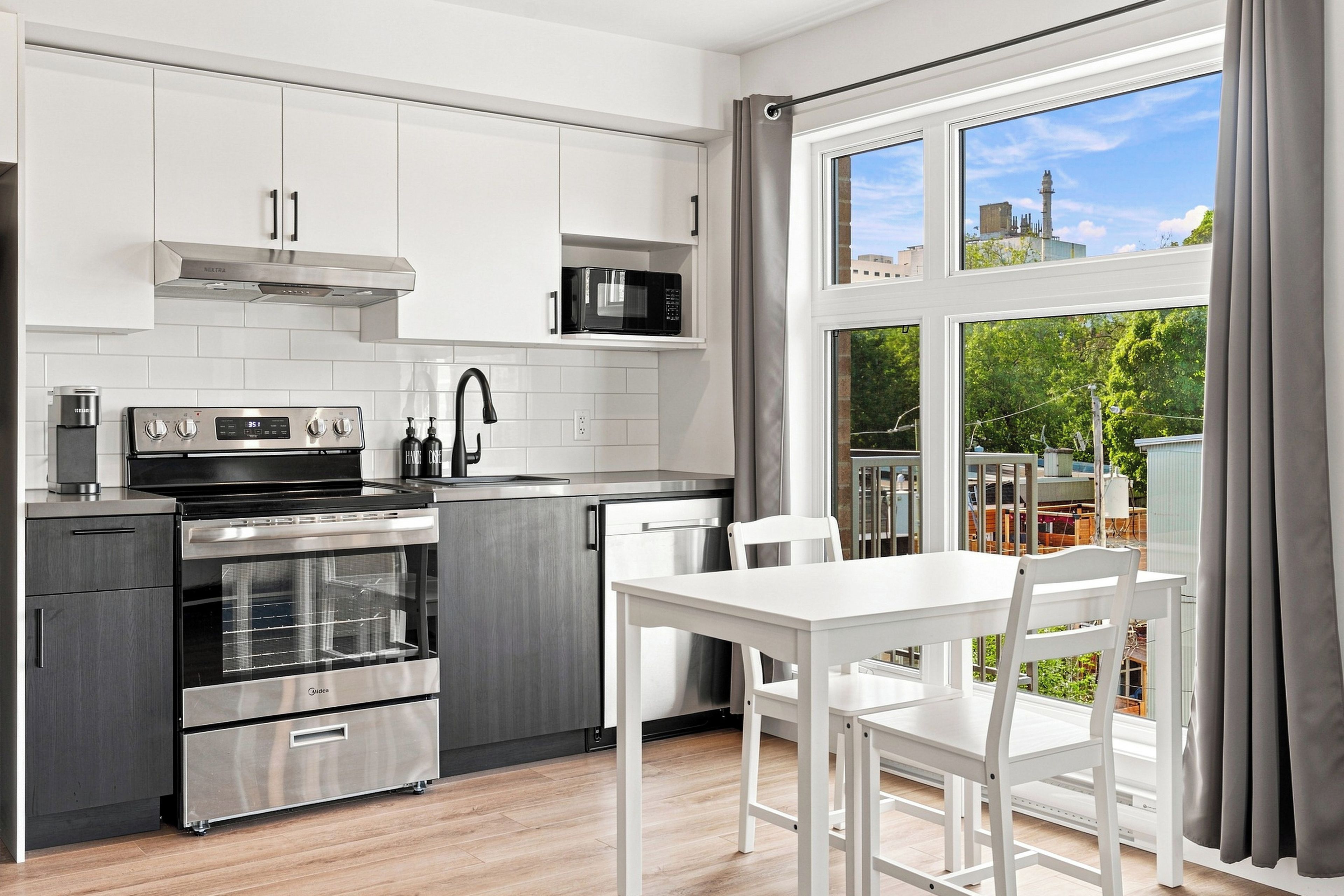 Una cocina moderna con electrodomésticos de acero inoxidable, un comedor con mesa y sillas blancas, y un amplio ventanal con vistas a edificios y vegetación.