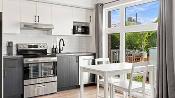 Una cocina moderna con electrodomésticos de acero inoxidable, un comedor con mesa y sillas blancas, y un amplio ventanal con vistas a edificios y vegetación.