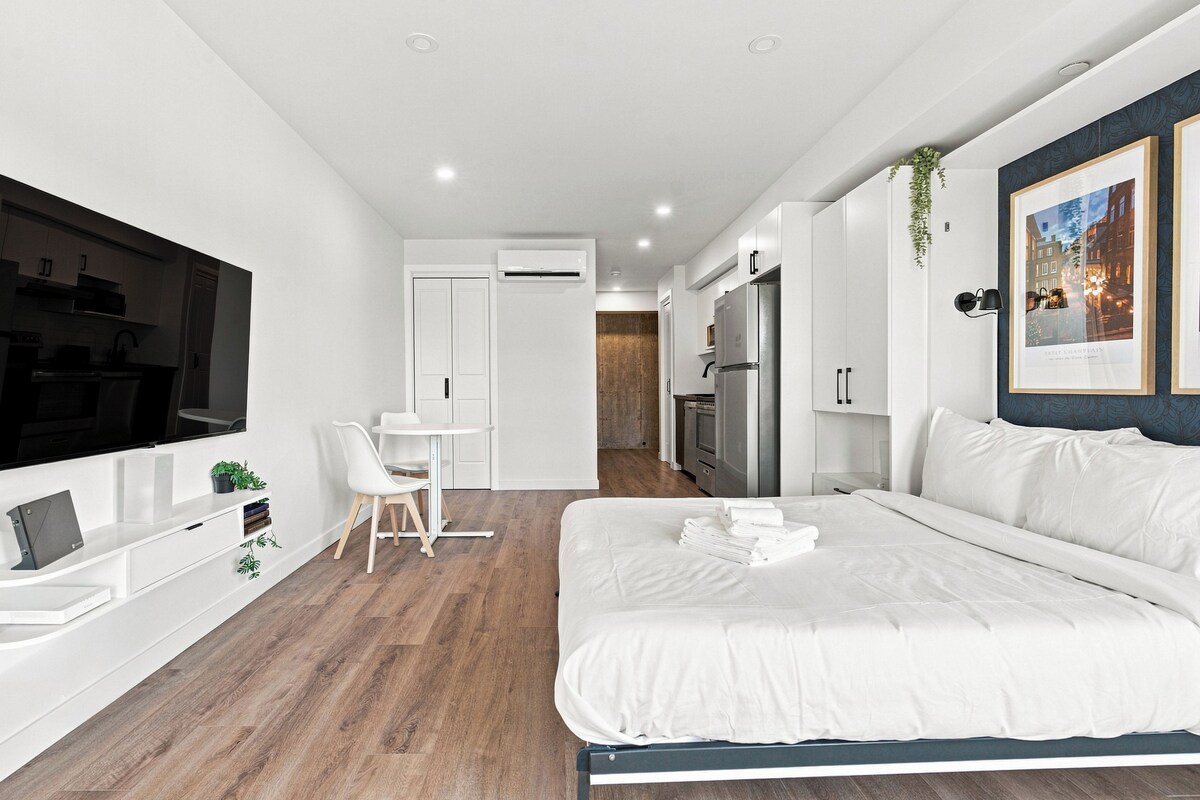 A modern bedroom with a bed, a desk, a TV, and a kitchenette.