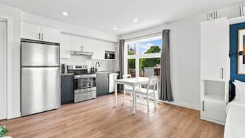 A modern kitchen with stainless steel appliances, a dining area with a white table and chairs, and a large window with a view of greenery.