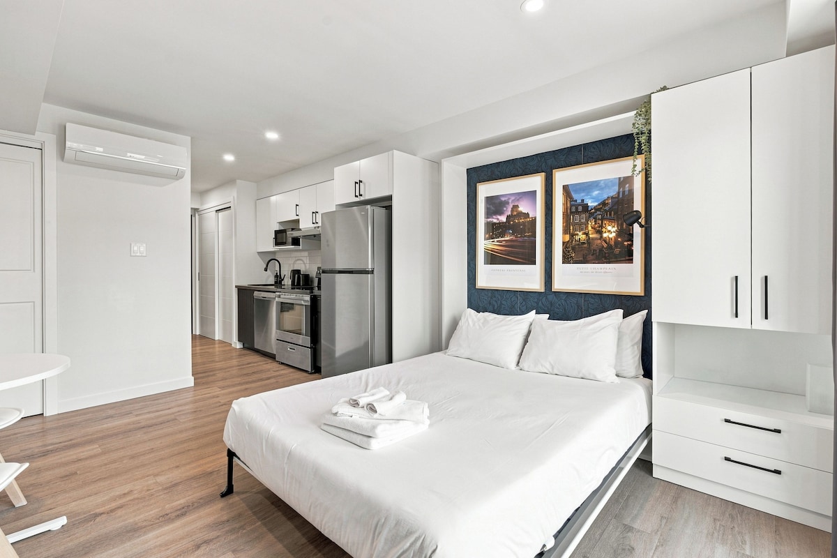 A modern hotel room with a large bed, a kitchenette, and a view of the city.