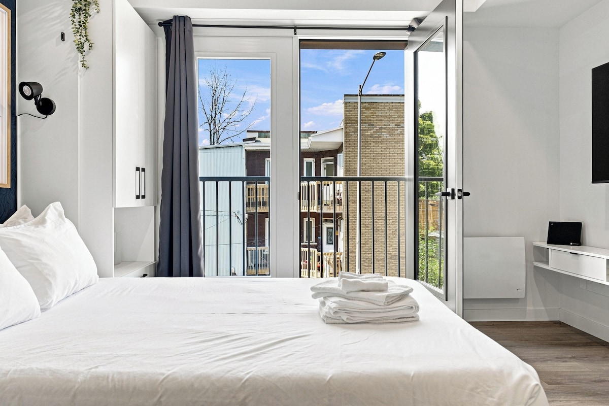 A modern bedroom with a large bed, a view to the outside, a small balcony, and a mounted security camera.