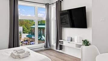 A hotel room with a bed, a TV mounted on the wall, a small desk with a laptop, and a view of the outside.