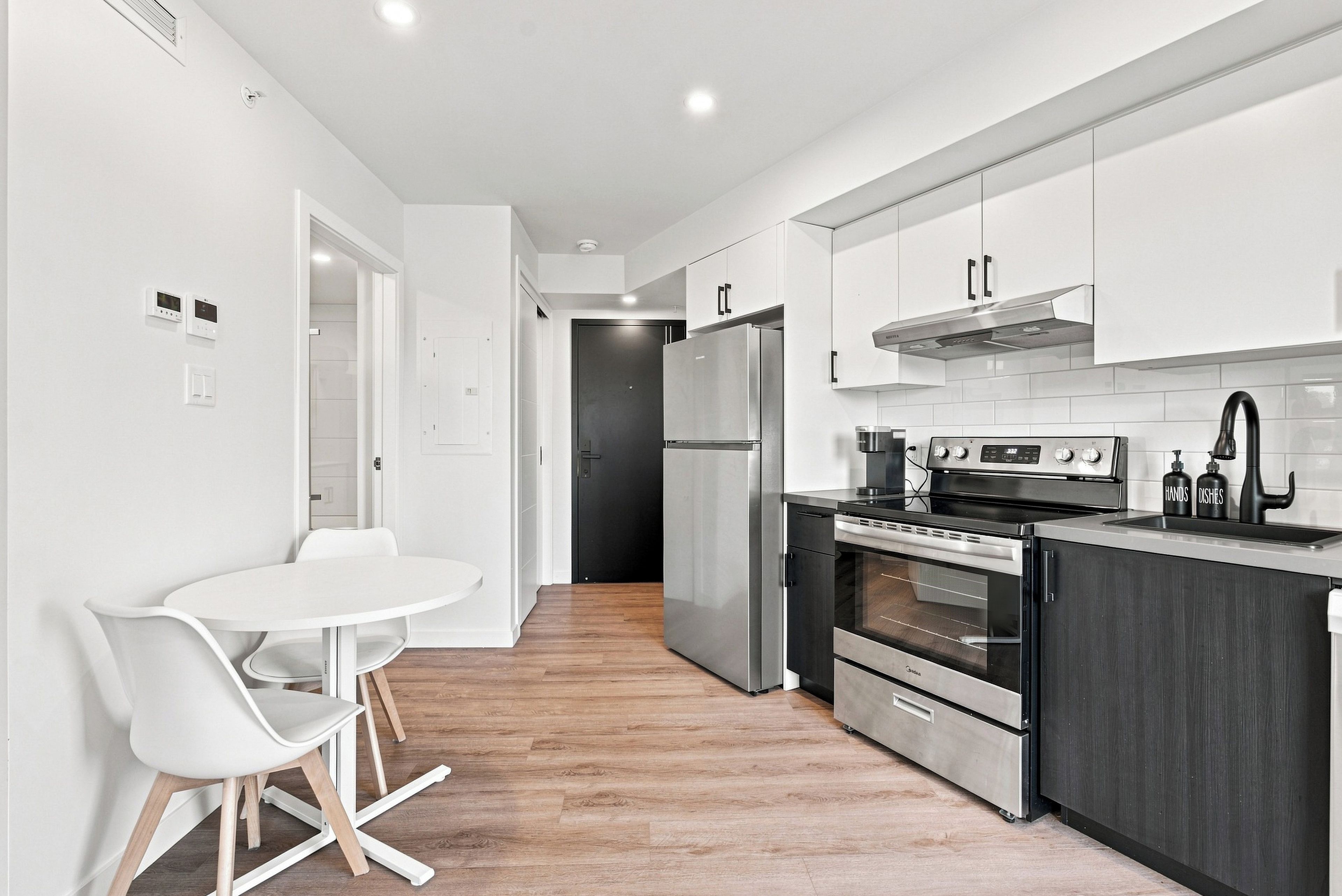 A modern kitchen with white cabinetry, a stainless steel refrigerator, an oven, and a microwave. There is a white dining table with chairs.