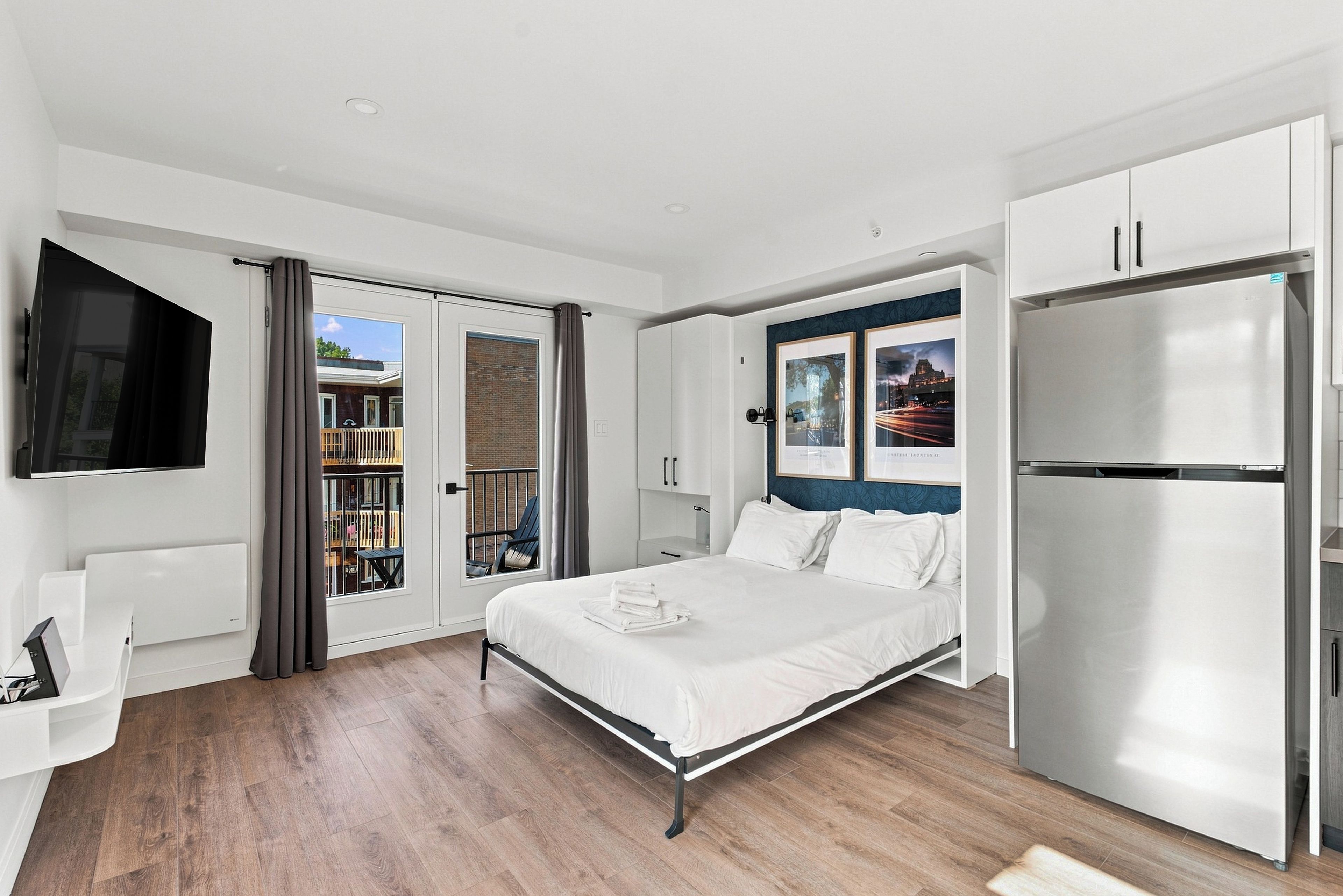 A modern bedroom with a bed, a flat-screen TV, a refrigerator, and a balcony with a view.