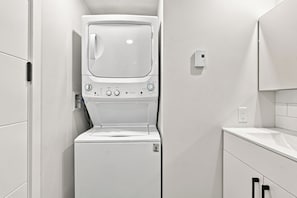 A compact, white laundry room with a front-loading washing machine and dryer, a small countertop, and a wall-mounted electrical outlet. - Appart'hôtel Viveo - Par Aneyro (Québec)