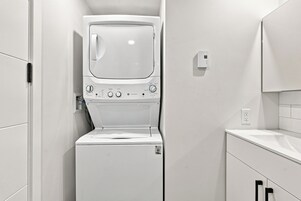 Une petite buanderie blanche, équipée d’un lave-linge et d’un sèche-linge à chargement frontal, d’un petit plan de travail et d’une prise électrique murale.