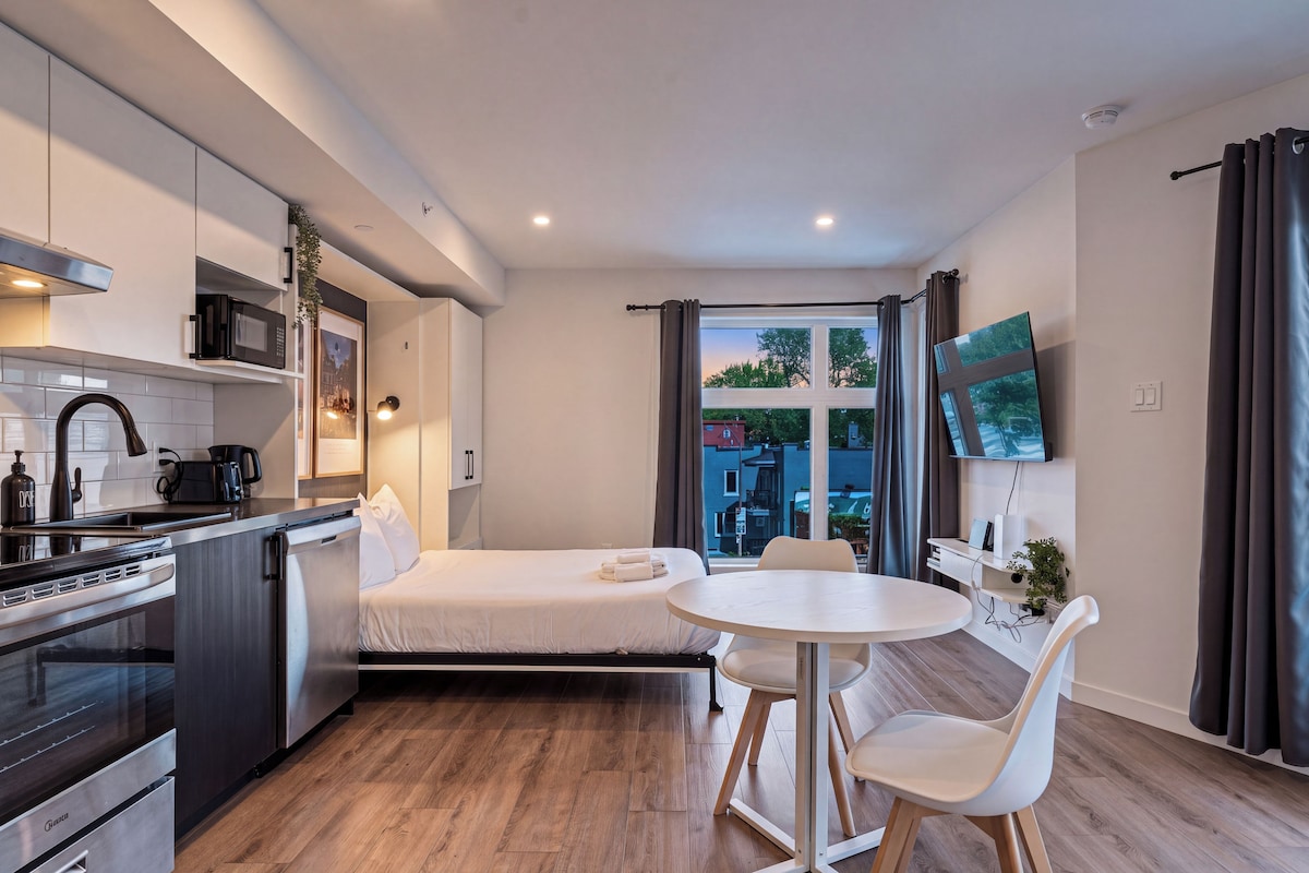 A compact hotel room with a kitchenette, a bed, a dining area, and a TV mounted on the wall.