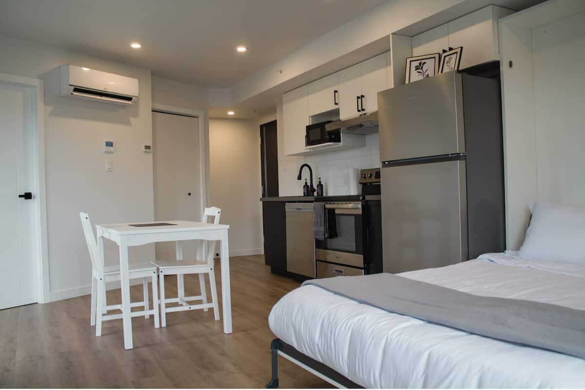 A modern bedroom with a bed, a small dining table with chairs, a kitchenette with a refrigerator, oven, and microwave.