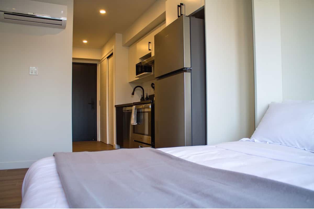 A bedroom with a bed, a kitchenette with a refrigerator, oven, and microwave, and an air conditioning unit.