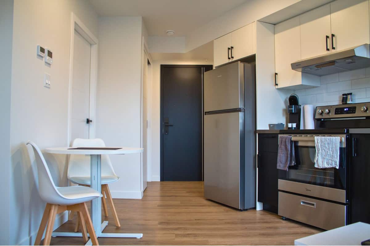 A modern kitchen with a stainless steel refrigerator, oven, and coffee maker. There is a small dining table with two chairs.