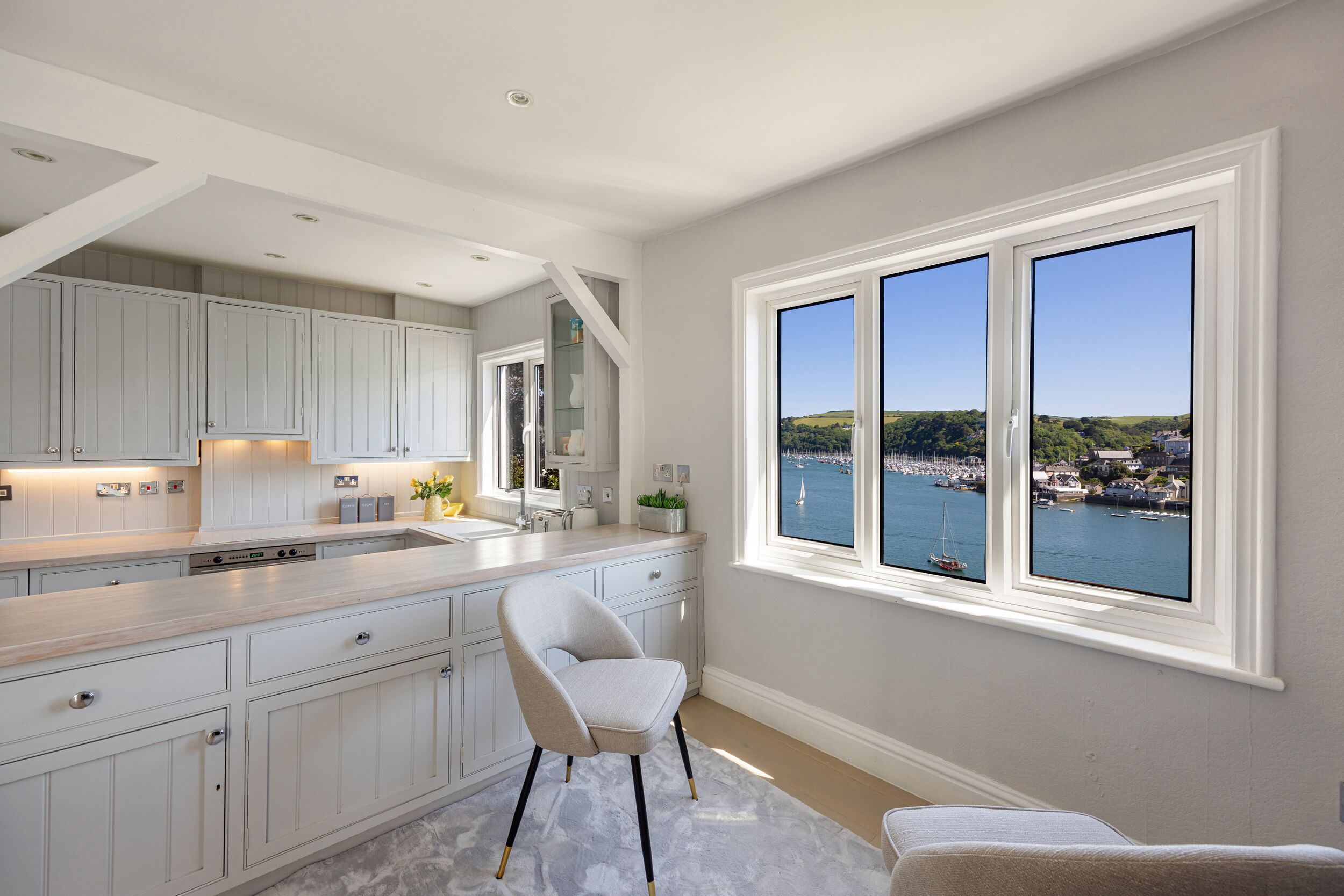 A modern kitchen with a view of a harbor, featuring white cabinetry, a marble countertop, and a large window.