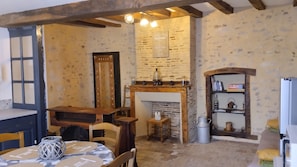 Un intérieur rustique avec des murs en briques apparentes, une table en bois recouverte d’une nappe à motifs, une cheminée avec un manteau de cheminée et une étagère remplie de livres et d’objets décoratifs.