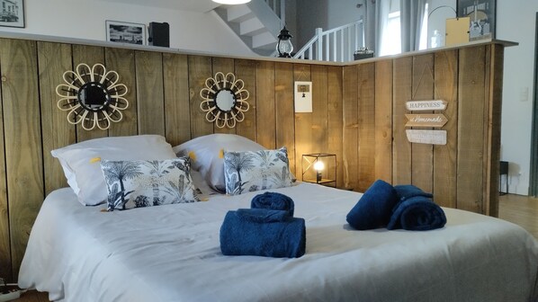 Une chambre avec une tête de lit en bois, deux horloges fixées au mur, un lit avec une literie blanche et un panneau indiquant : « Le BONHEUR se crée chez soi ».