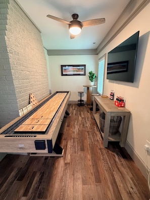 Una habitación con mesa de juego de paletas, ventilador de techo, televisor montado y una mesita con botellas.