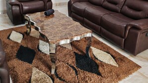 Una sala de estar con un sofá de cuero marrón, un sillón de cuero marrón, una mesa de centro de mármol, un televisor en la pared y una alfombra estampada.