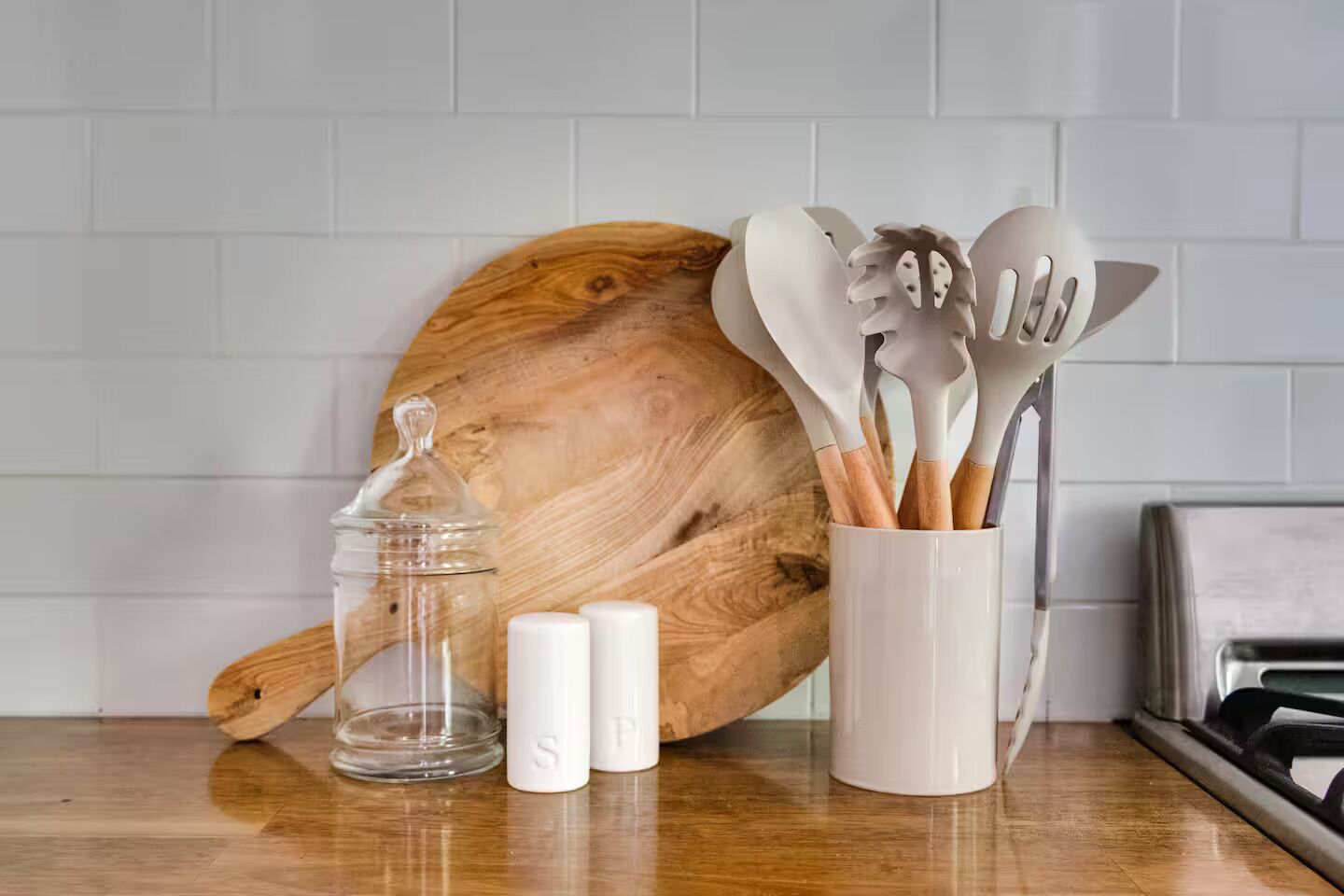 A kitchen counter with a wooden cutting board, a glass jar, salt shakers, and a utensil holder.