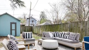 Un patio con un divano angolare grigio, due poltrone, un poggiapiedi rotondo e cuscini decorati.