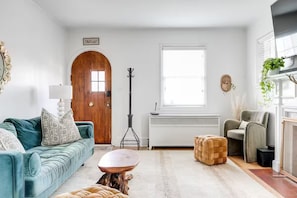 A living room with a teal sofa, a coffee table, a woven ottoman, a wooden door, a standing lamp, a television, and a window. - Relaxing & Quiet Retreat Fully Fenced Backyard (Richmond)