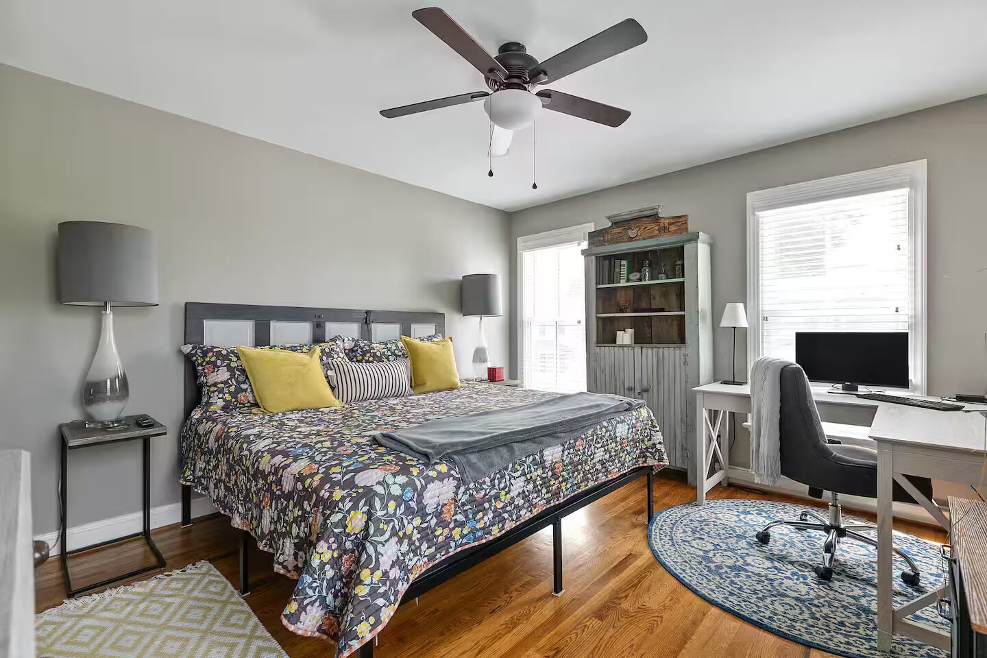 A bedroom with a bed, a desk, a chair, a ceiling fan, a bookshelf, and a window.