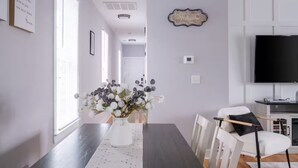 Et spiseområde med et langt bord, hvide stole, en vase med blomster og et fjernsyn på væggen.