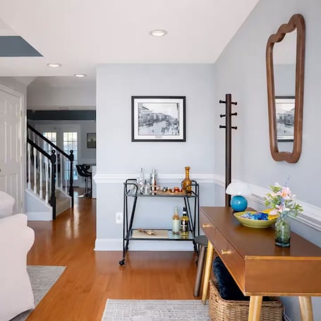 A modern living room with a wooden dining table, a black metal side table, a framed black and white photograph on the wall, and a large mirror.